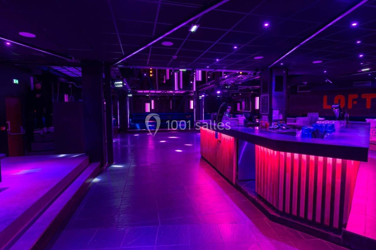 Salle de discothèque vide avec éclairage violet et rose, bar illuminé et piste de danse en arrière-plan.
