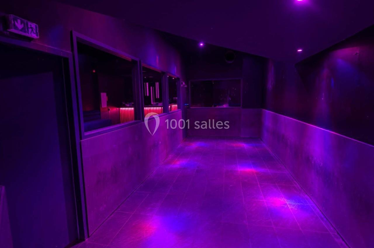 Couloir sombre éclairé par des lumières violettes, menant à une salle avec un comptoir lumineux en arrière-plan.