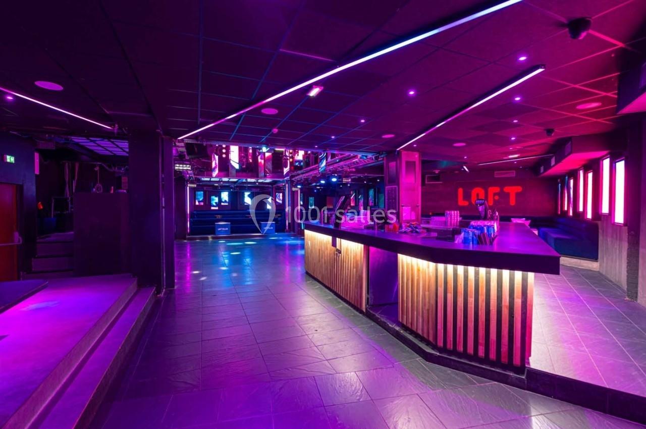 Salle de discothèque moderne avec éclairage violet, bar en bois éclairé et piste de danse vide.