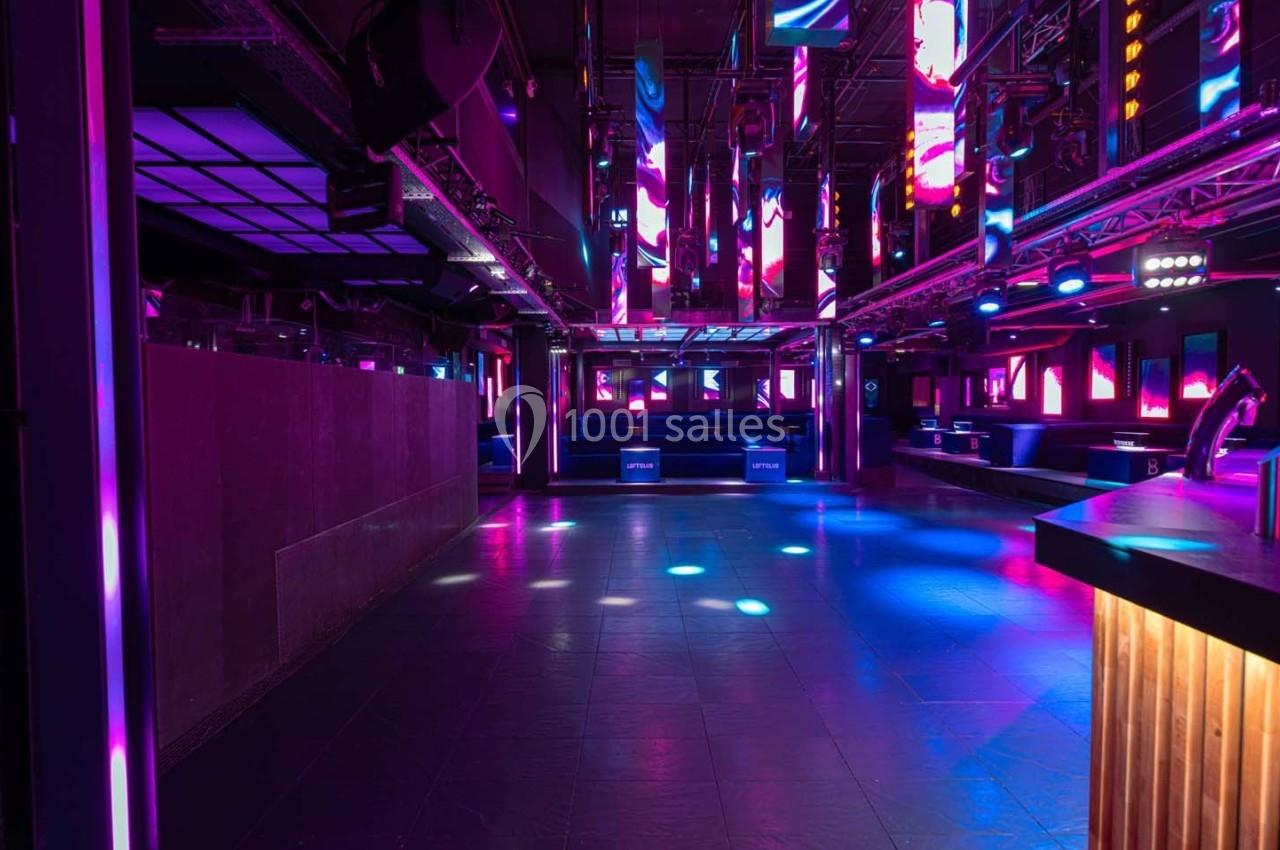 Salle de discothèque vide avec éclairage coloré et néons, piste de danse au centre et bar en bois à droite.