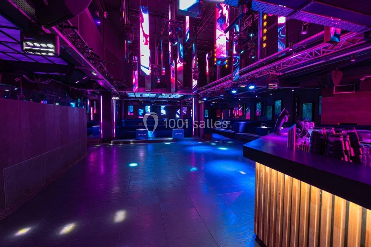 Salle de discothèque vide avec éclairage néon coloré, piste de danse centrale et bar en bois à droite.