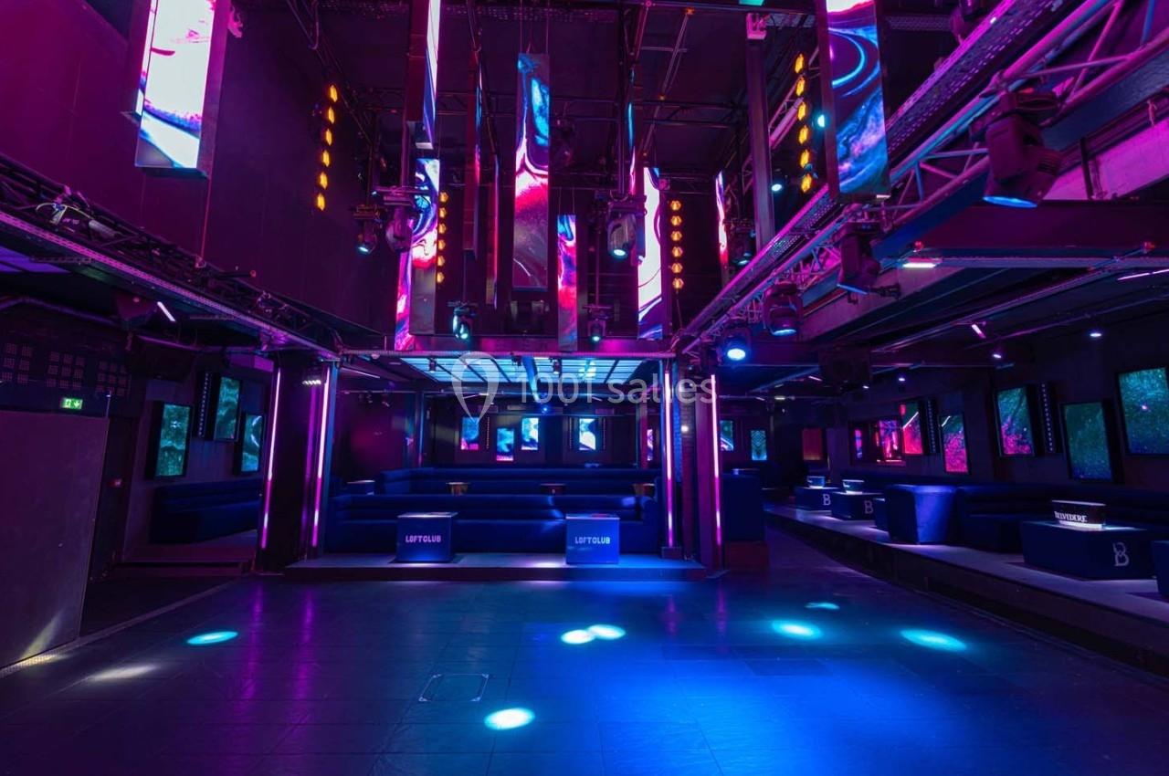 Salle de discothèque vide avec éclairage néon coloré, écrans lumineux et banquettes alignées le long des murs.