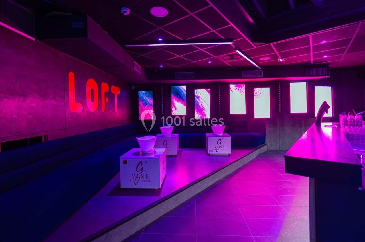 Salle de club avec éclairage rose, banquettes bleues, tables basses et écrans lumineux au mur.