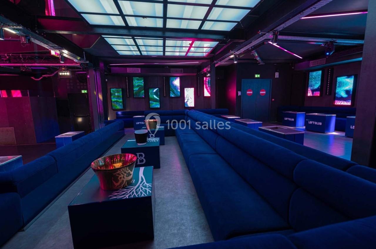 Salle de club moderne avec banquettes bleues, tables basses et éclairage néon rose et bleu.