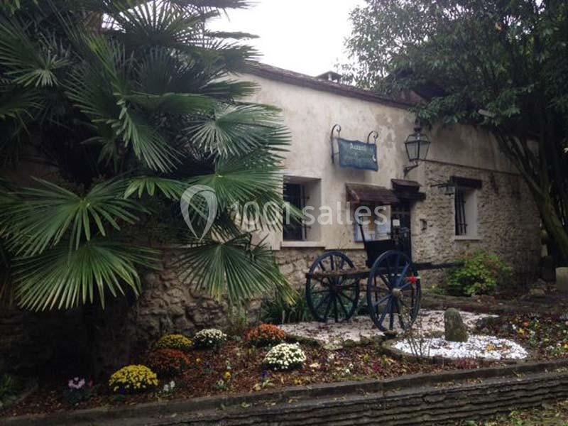 Façade en pierre d'un bâtiment ancien avec une charrette en bois, entourée de végétation et de fleurs.