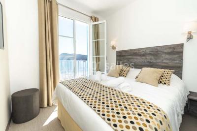 Chambre lumineuse avec lit double, décor beige et fenêtre ouverte offrant une vue sur la mer et les collines.