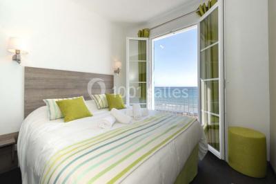 Chambre lumineuse avec lit double, décor beige et fenêtre ouverte offrant une vue sur la mer et les collines.