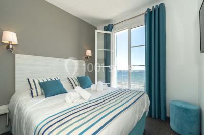 Chambre lumineuse avec lit double, décor beige et fenêtre ouverte offrant une vue sur la mer et les collines.