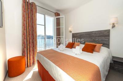 Chambre lumineuse avec lit double, décor beige et fenêtre ouverte offrant une vue sur la mer et les collines.