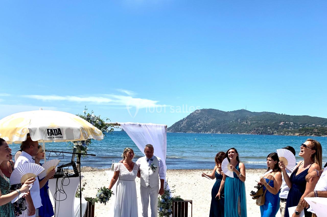 Cérémonie de mariage sur une plage, avec un couple sous une arche blanche entouré d'invités et vue sur la mer.