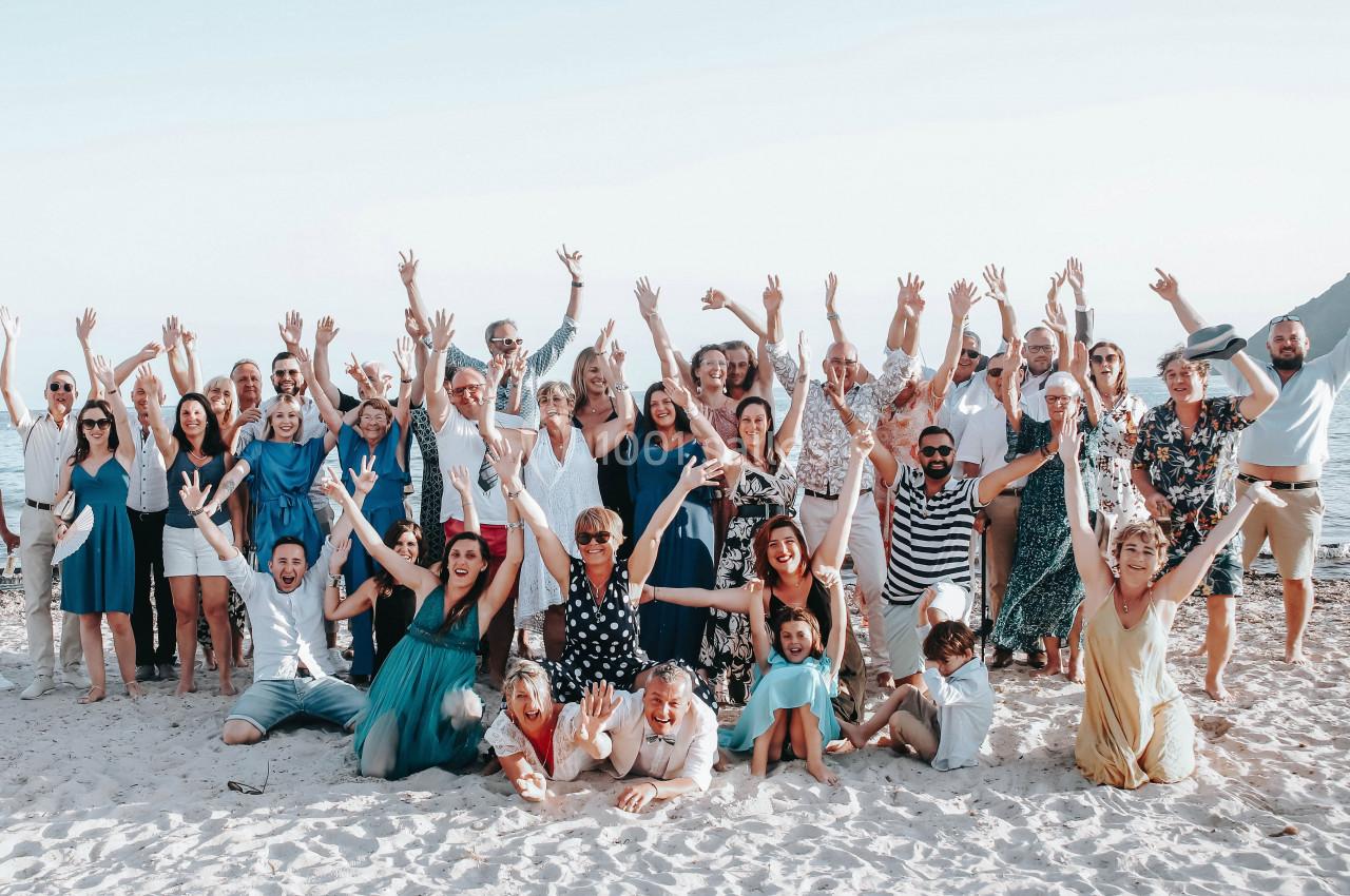 Un groupe de personnes souriantes posant sur une plage, bras levés, avec la mer et des collines en arrière-plan.