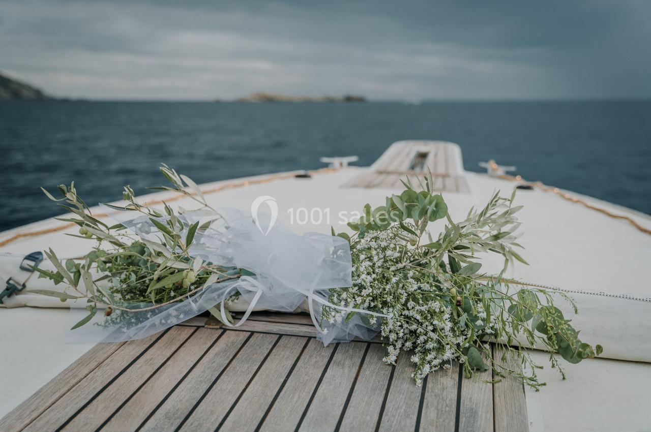 Bouquet de fleurs blanches et feuillages verts posé sur le pont en bois d'un bateau, avec la mer en arrière-plan.