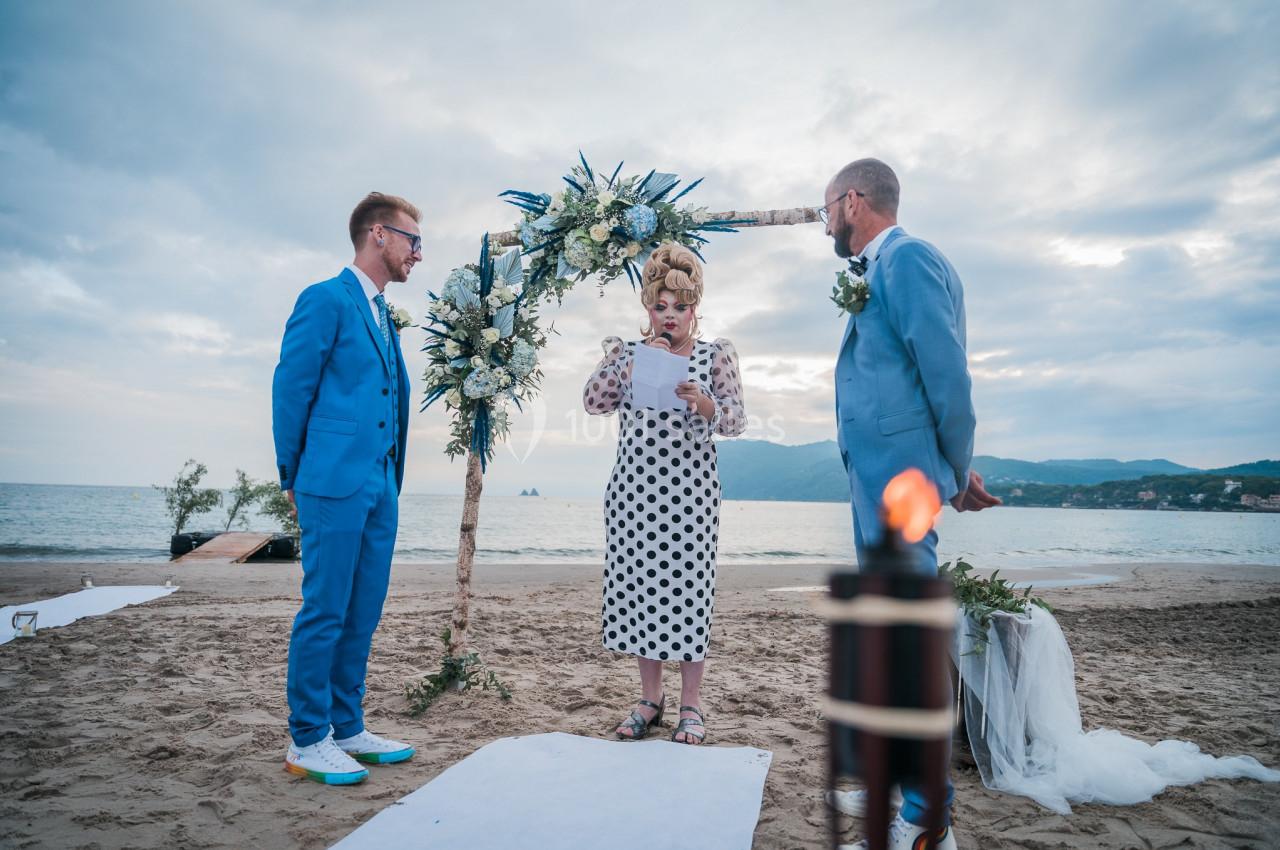 Deux hommes en costume assistent à une cérémonie de mariage sur une plage, officiée par une femme lisant un texte.
