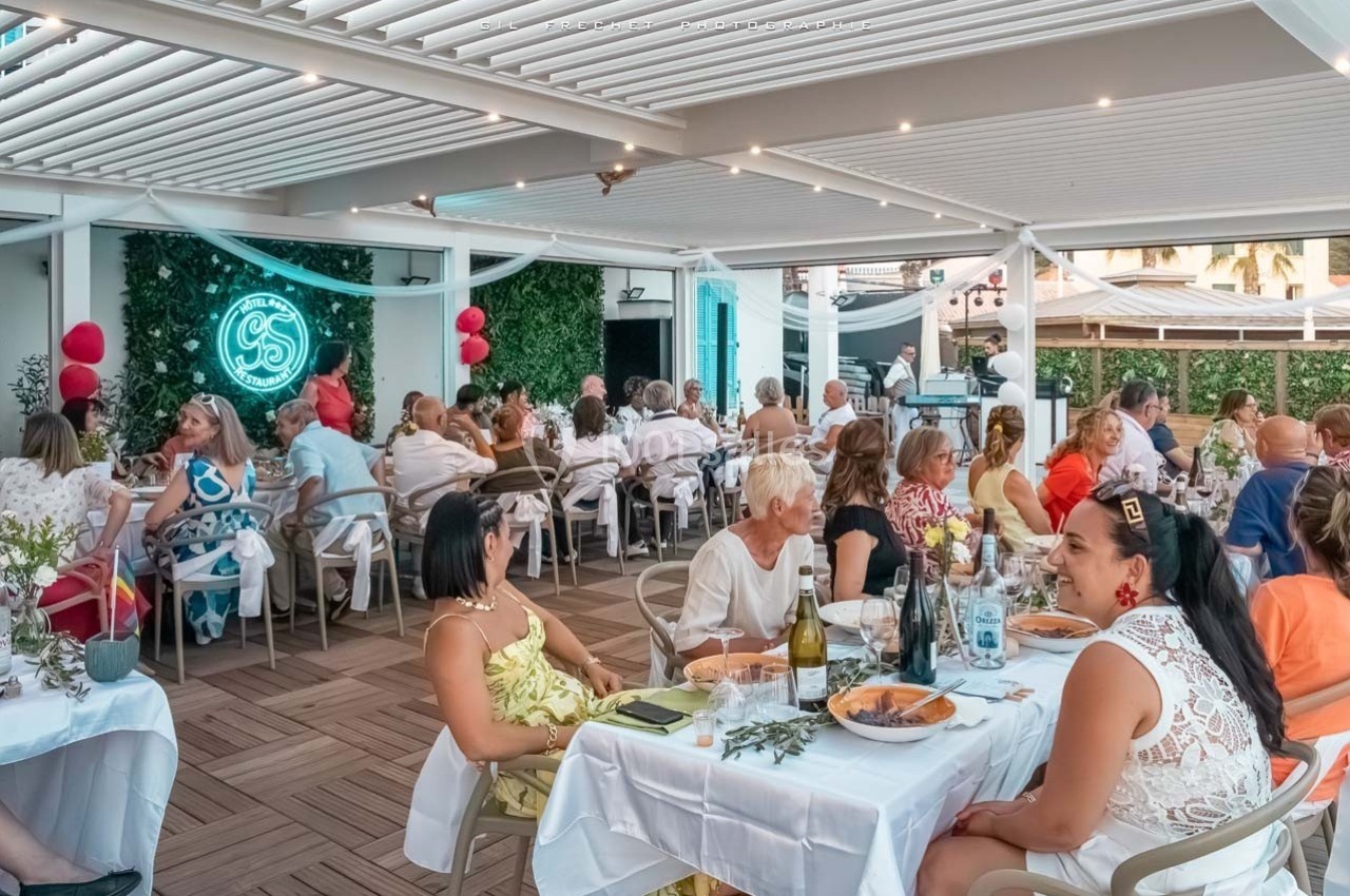 Des convives dînent en terrasse couverte, décorée de plantes et d'un néon lumineux, dans une ambiance conviviale.