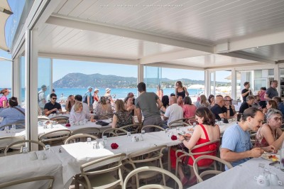 Chambre lumineuse avec lit double, décor beige et fenêtre ouverte offrant une vue sur la mer et les collines.