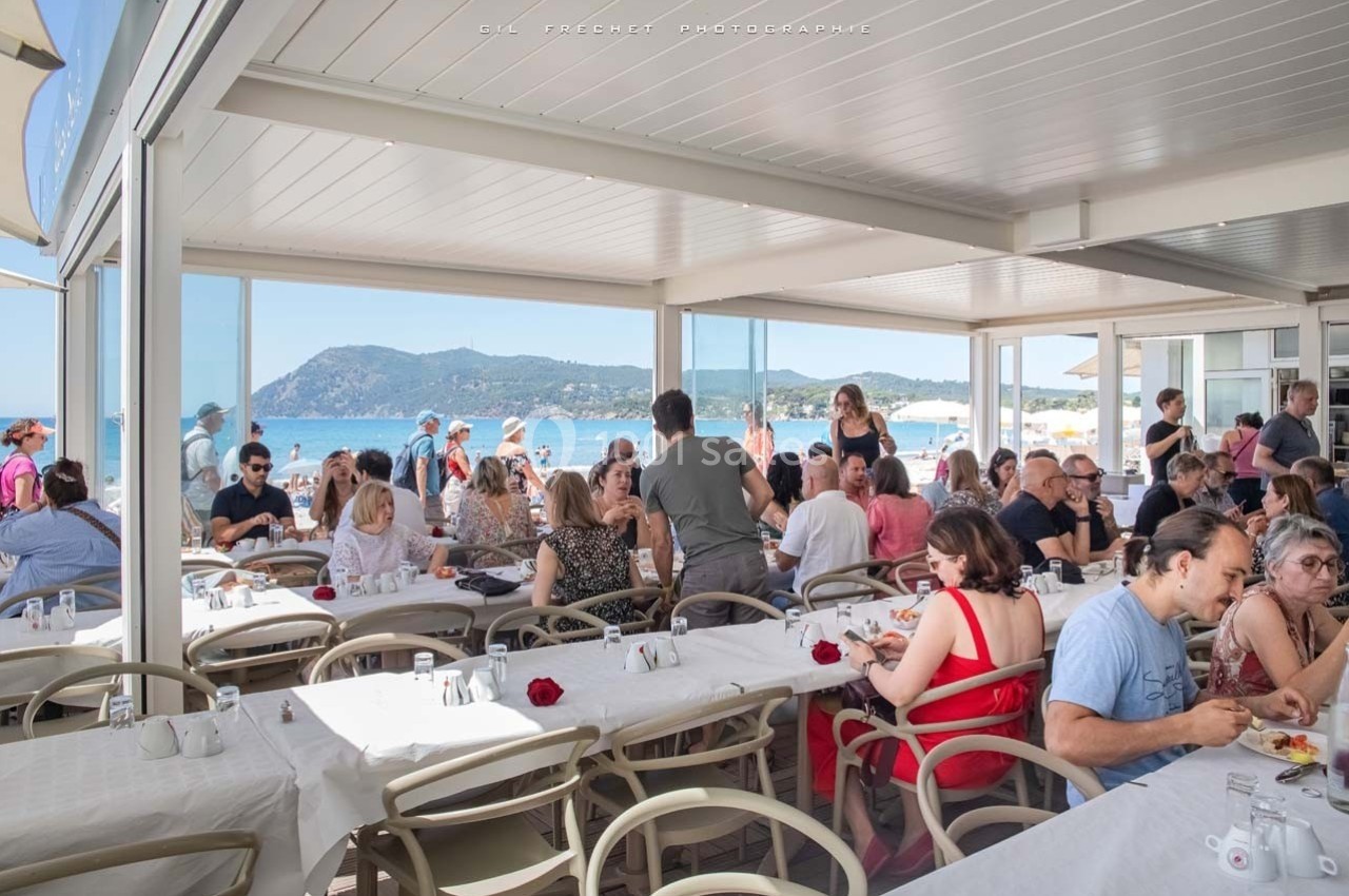 Salle de restaurant en bord de mer avec des clients attablés, vue sur la plage et les montagnes en arrière-plan.