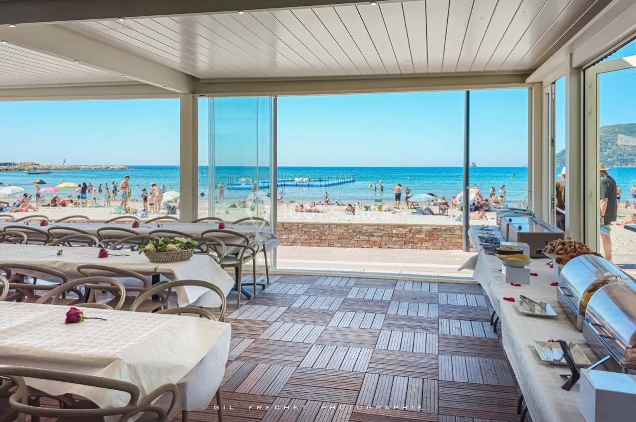 Salle de restaurant ouverte sur une plage ensoleillée avec vue sur la mer et des baigneurs au loin.