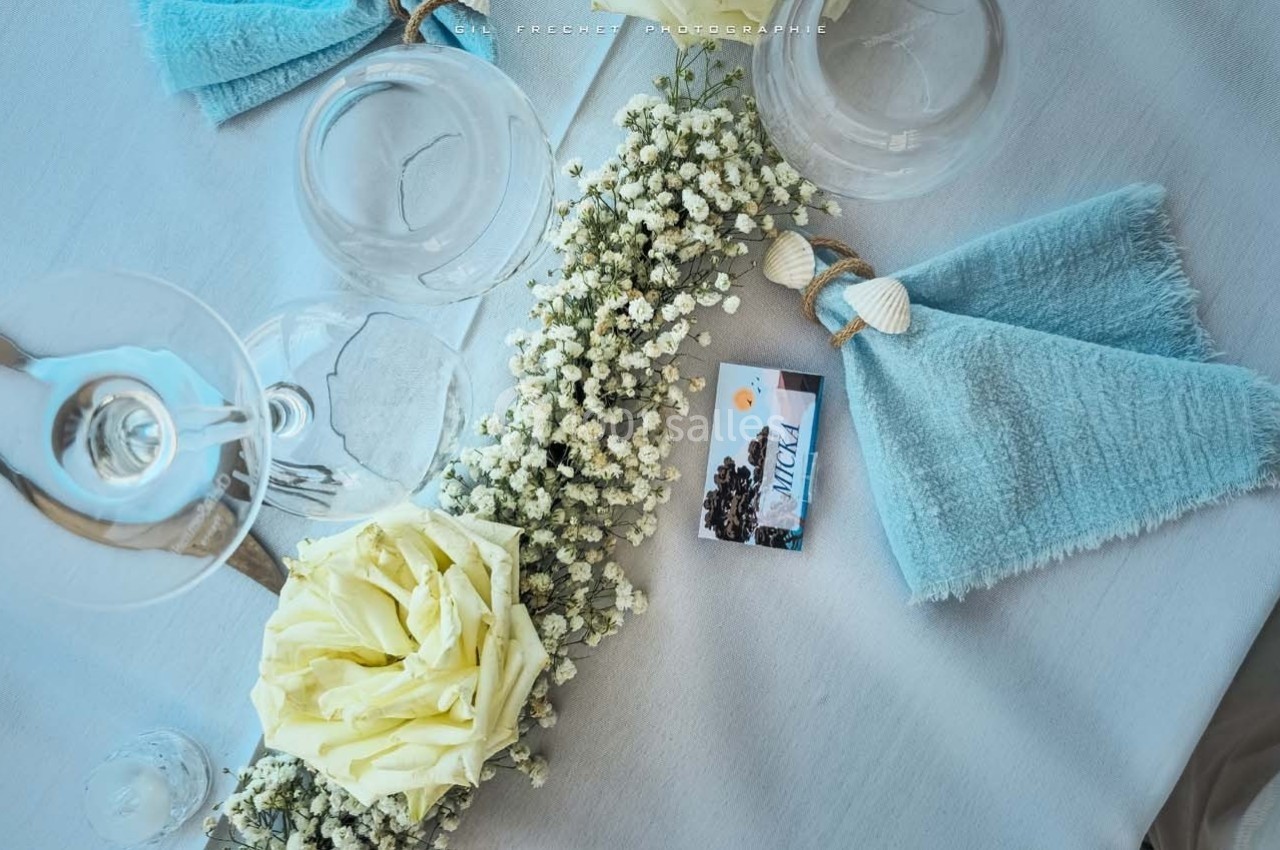 Serviette bleue pliée avec un nœud, rose blanche et gypsophile sur une table dressée avec des verres.