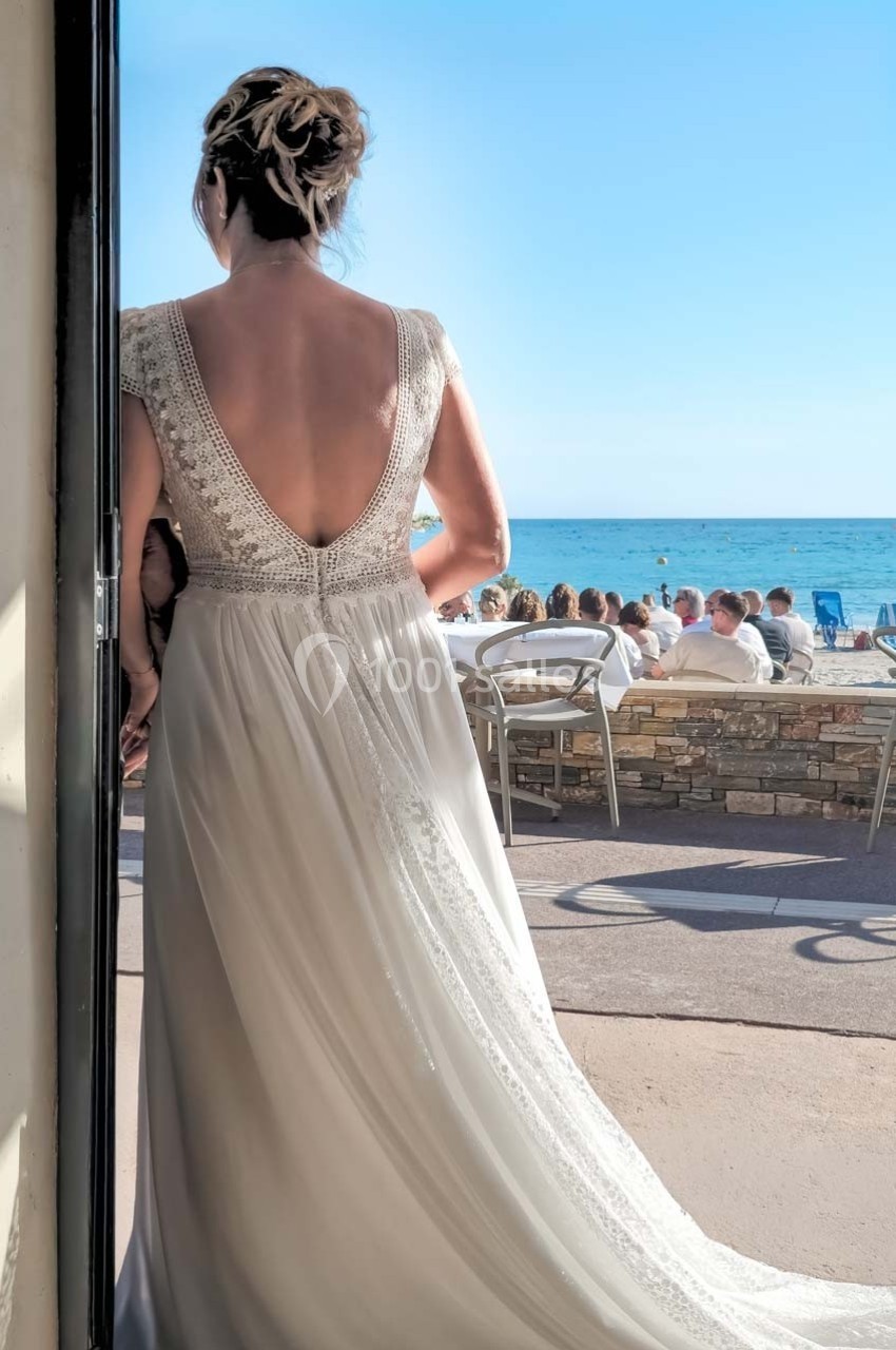 Une femme en robe de mariée dos à la caméra, face à une terrasse en bord de mer par une journée ensoleillée.