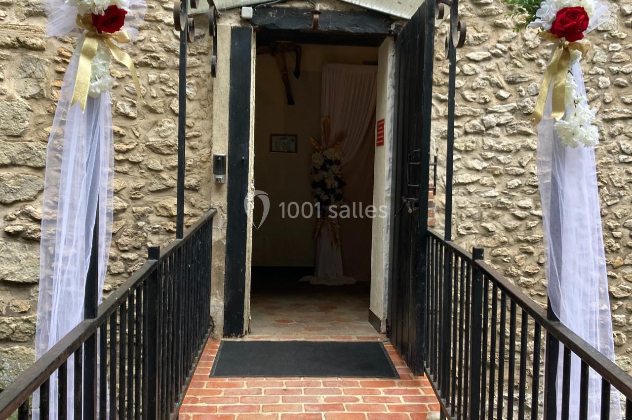 Entrée en briques avec une arche métallique décorée de fleurs blanches et rouges menant à une porte ouverte.