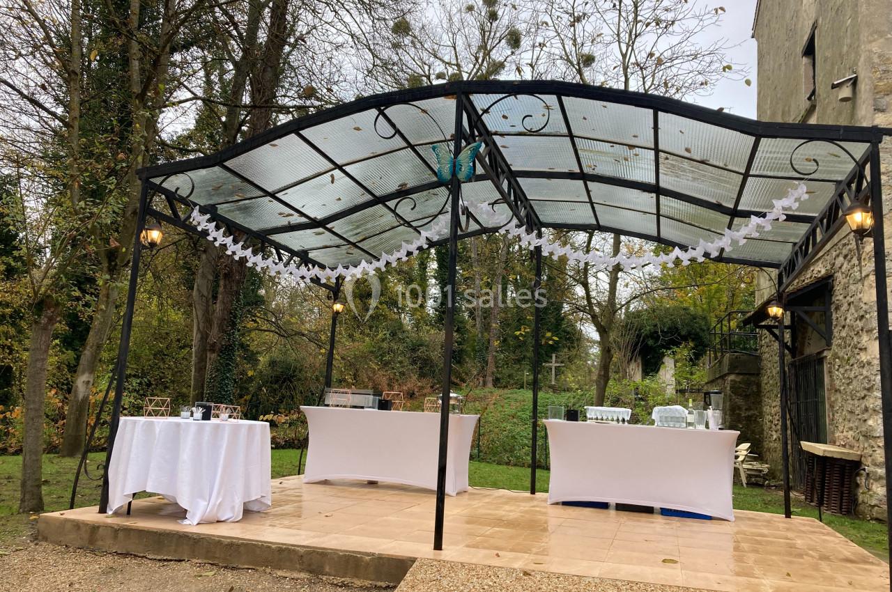 Tonnelle en métal avec toit transparent, décorée de guirlandes blanches, entourée de verdure dans un jardin.