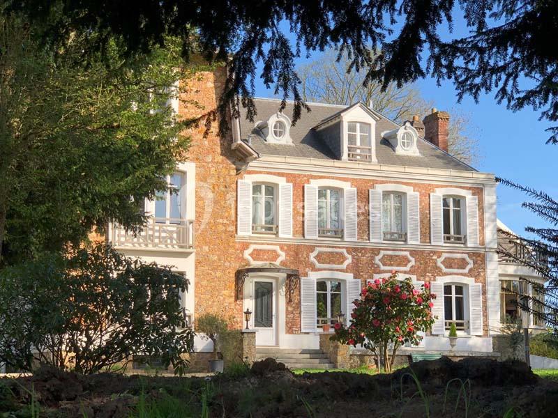 Façade d'une maison en briques rouges avec volets blancs, entourée de végétation et éclairée par le soleil.
