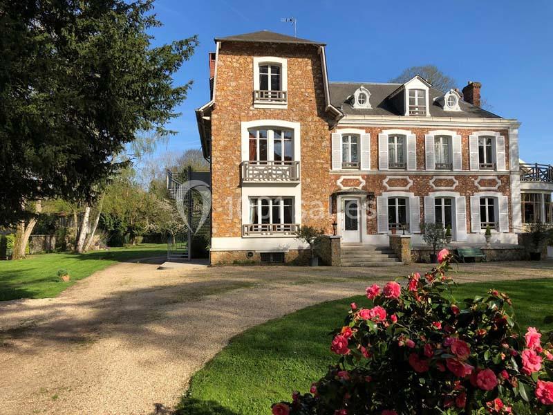 Grande maison en briques et pierres avec jardin arboré, allée gravillonnée et massif de fleurs au premier plan.