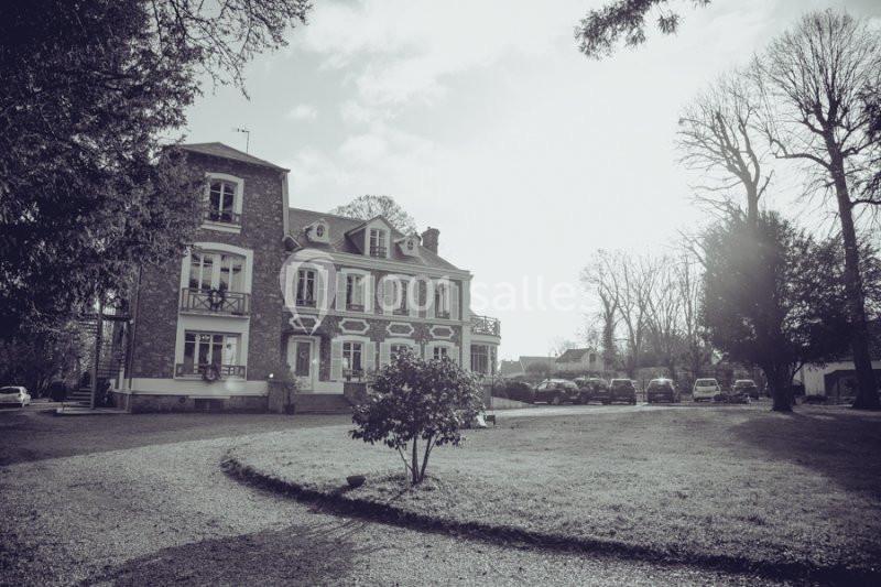 Façade d'une grande maison en pierre entourée d'un jardin avec des arbres, sous un ciel partiellement nuageux.