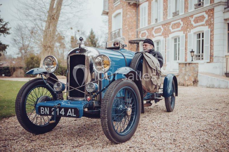 Voiture ancienne bleue stationnée devant une maison en briques, avec deux personnes assises à l'intérieur.