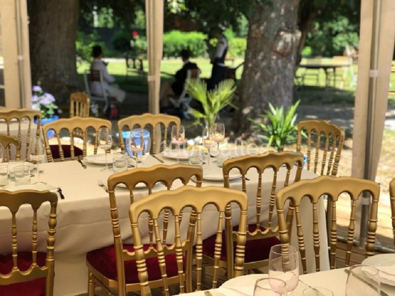 Salle de réception avec tables dressées, chaises dorées et vue sur un jardin arboré en arrière-plan.