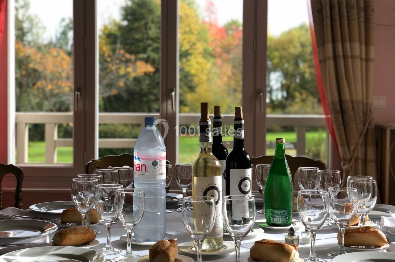 Table dressée avec des verres, bouteilles de vin, eau plate et gazeuse, et petits pains, devant une fenêtre donnant sur un…