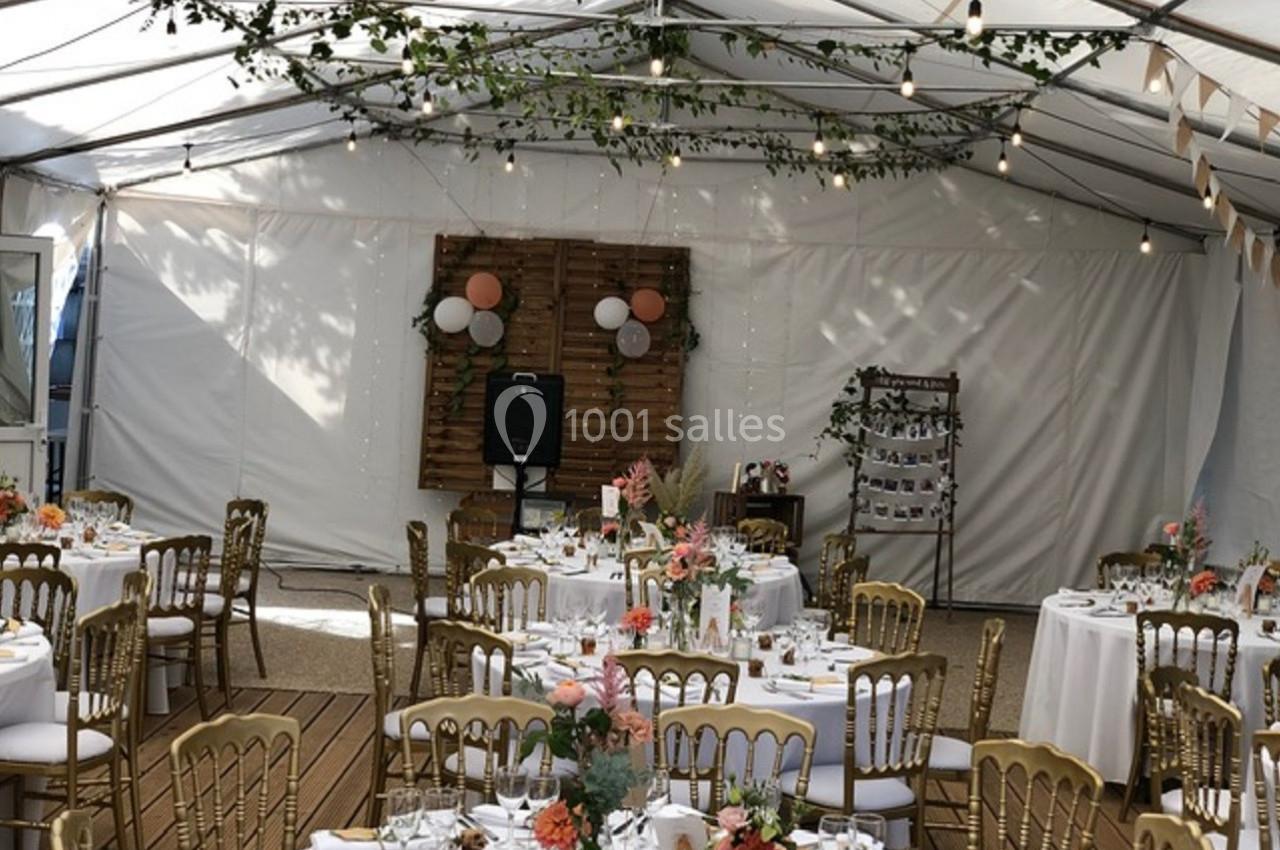 Salle de réception sous tente avec tables décorées, chaises dorées, guirlandes lumineuses et arrangements floraux.