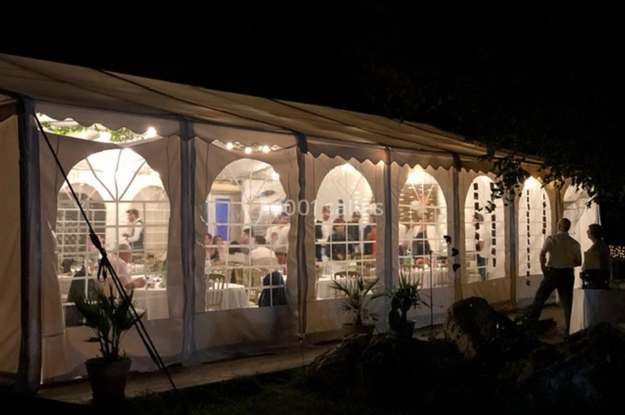 Vue nocturne d'une tente éclairée où des personnes participent à un événement, entourée de plantes et de rochers.