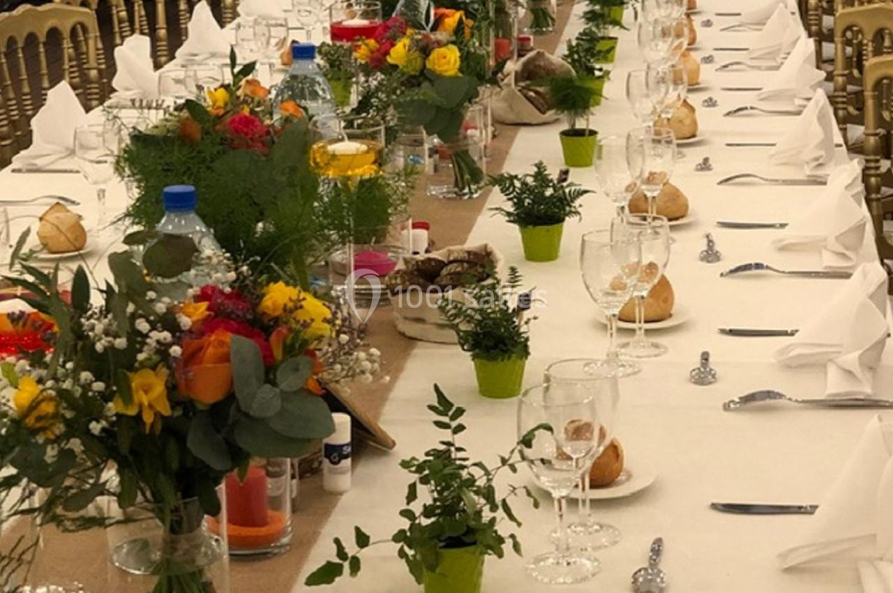 Table de banquet dressée avec nappes blanches, verres, serviettes pliées, fleurs colorées et petits pots de plantes.
