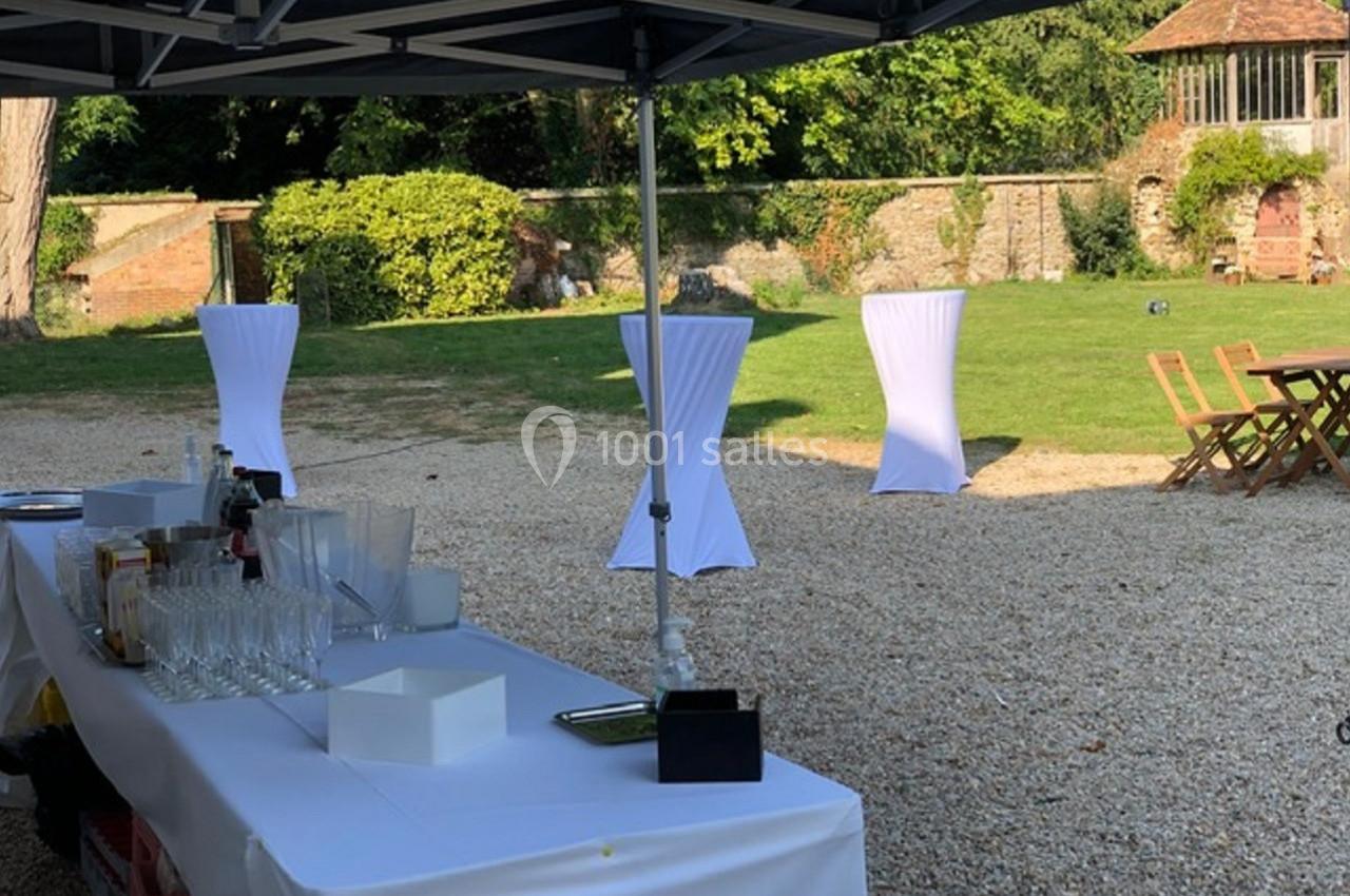 Table dressée sous un barnum avec verres et boissons, dans un jardin avec des tables hautes et un kiosque en arrière-plan.