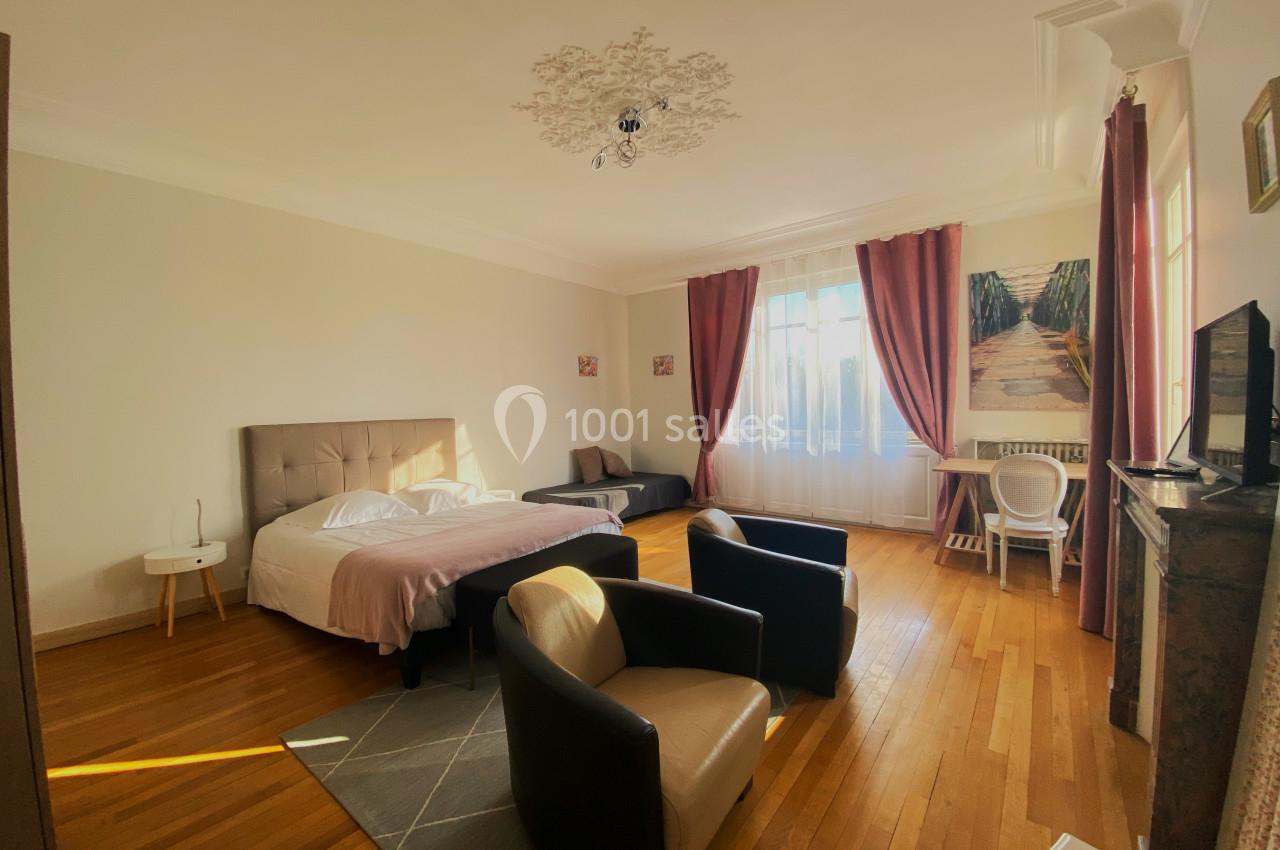 Chambre lumineuse avec lit double, fauteuils, bureau, téléviseur, parquet et grandes fenêtres avec rideaux.