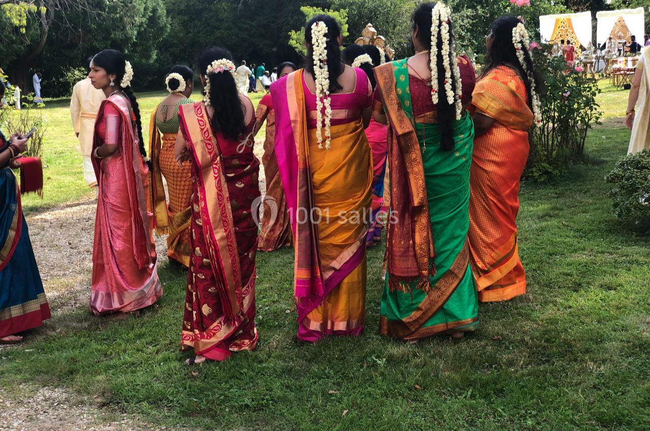 Des femmes en saris colorés avec des guirlandes de fleurs dans les cheveux, debout dans un jardin lors d'un événement.