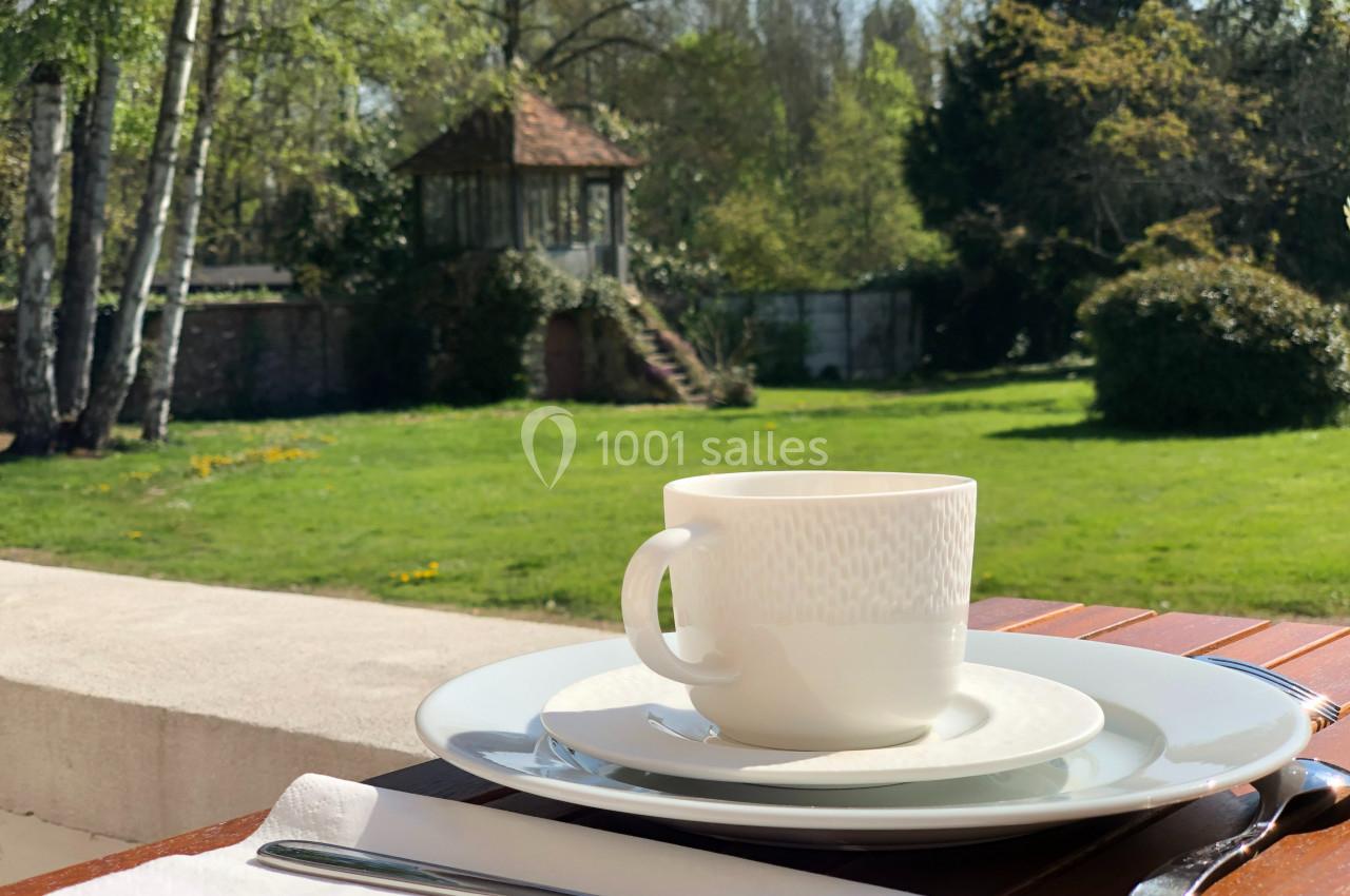 Tasse blanche posée sur une assiette, sur une table en bois, avec un jardin verdoyant en arrière-plan.