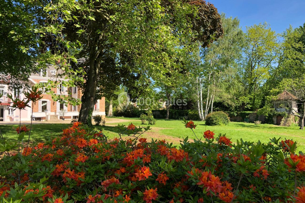 Jardin verdoyant avec des fleurs orange au premier plan, une maison en briques et des arbres en arrière-plan.