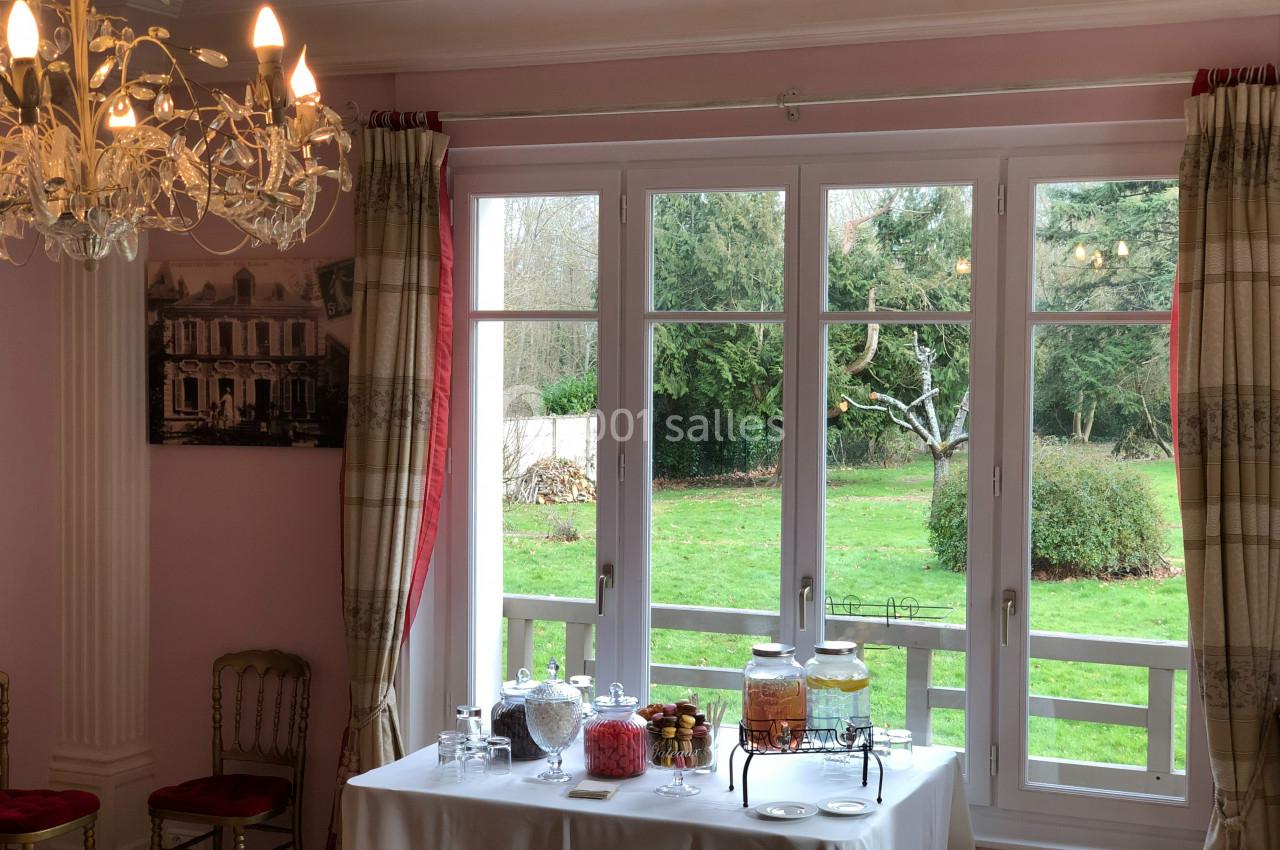Table dressée avec des boissons et des bonbonnières devant une grande fenêtre donnant sur un jardin verdoyant.