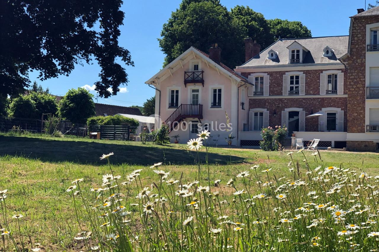 Maison avec façade claire entourée de verdure et de marguerites, située près d'autres bâtiments en pierre.