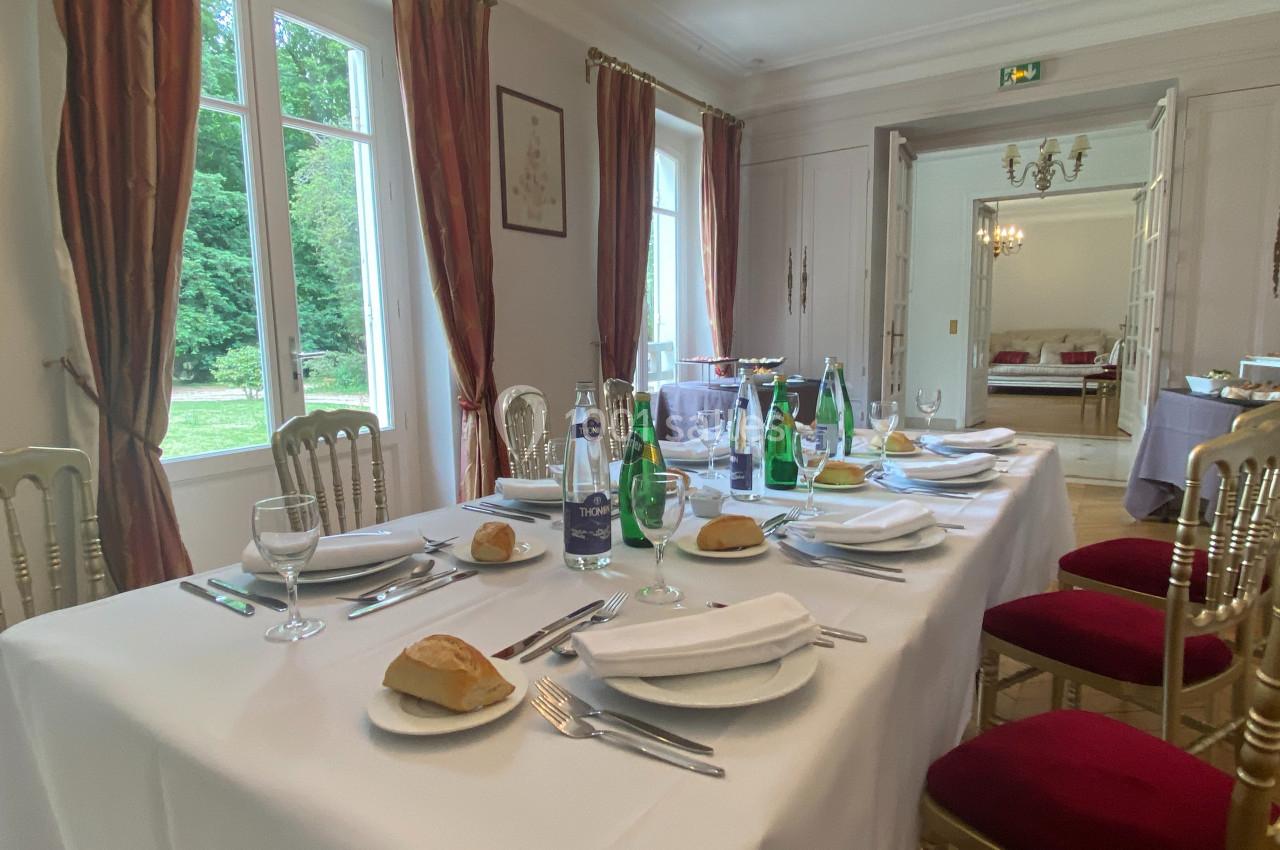 Table dressée avec nappes blanches, couverts, verres, bouteilles d'eau et pain, dans une salle lumineuse avec vue sur un…