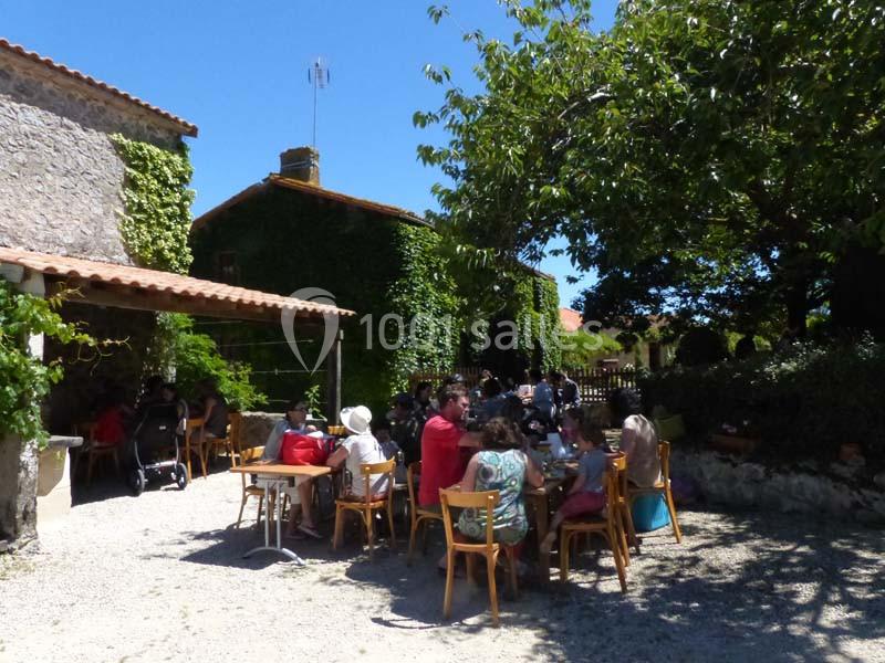 Des personnes assises à des tables en extérieur, près de bâtiments en pierre couverts de végétation, sous un ciel bleu.
