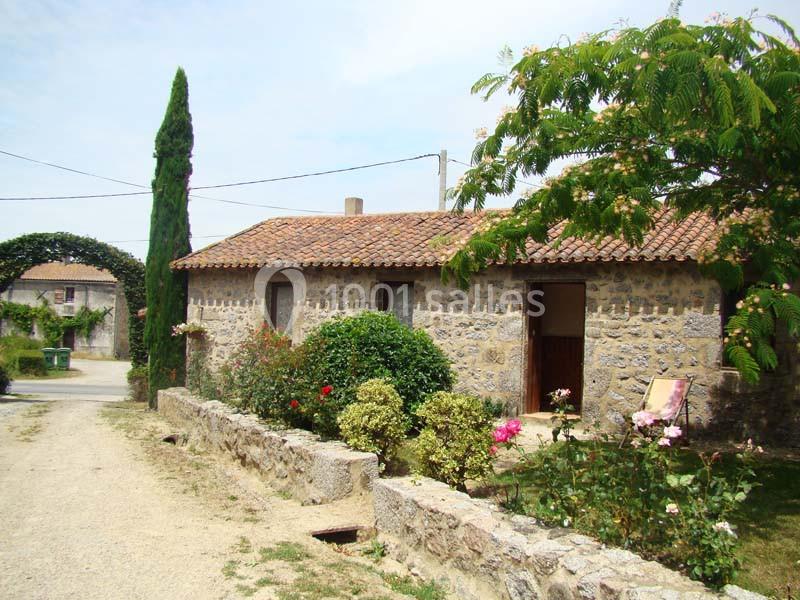 Maison en pierre avec toit en tuiles, jardin fleuri et allée bordée d'arbres dans un cadre rural.