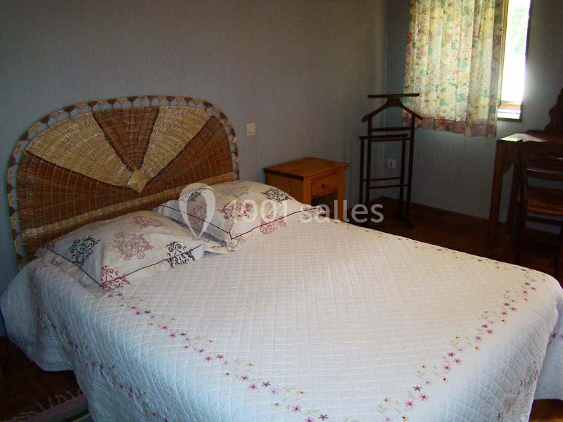 Lit double avec tête de lit en osier, drap blanc brodé, table de chevet en bois et fenêtre avec rideaux fleuris.