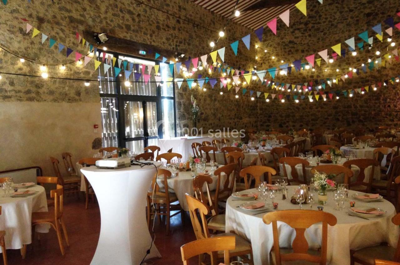 Salle décorée pour un événement avec guirlandes colorées, tables rondes dressées et murs en pierre apparente.