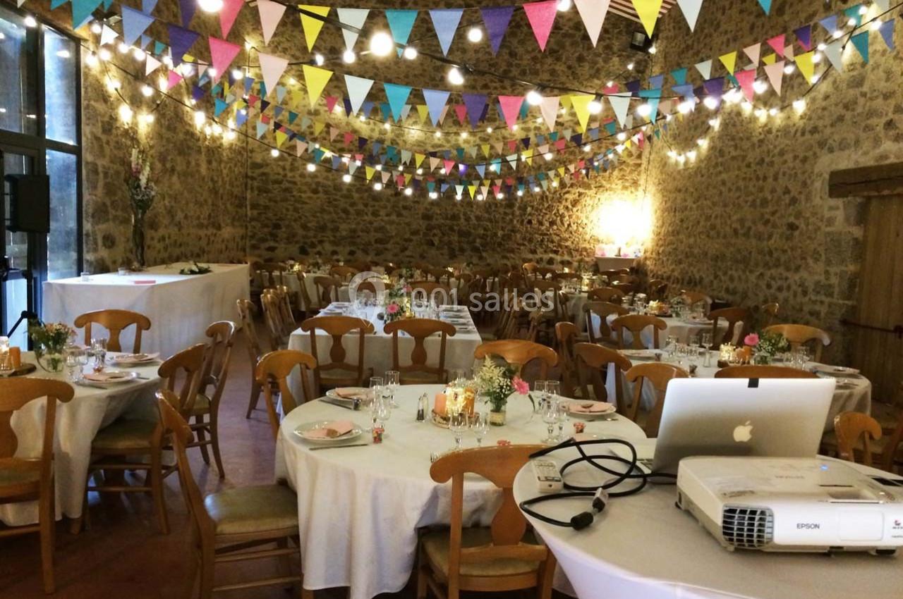 Salle de réception décorée de guirlandes colorées et éclairée, avec tables rondes dressées pour un événement.