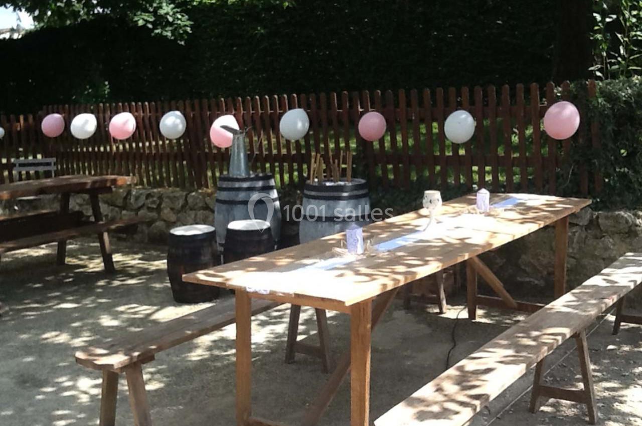 Tables en bois et bancs disposés en extérieur, décorés de ballons roses et blancs près d'une clôture en bois.