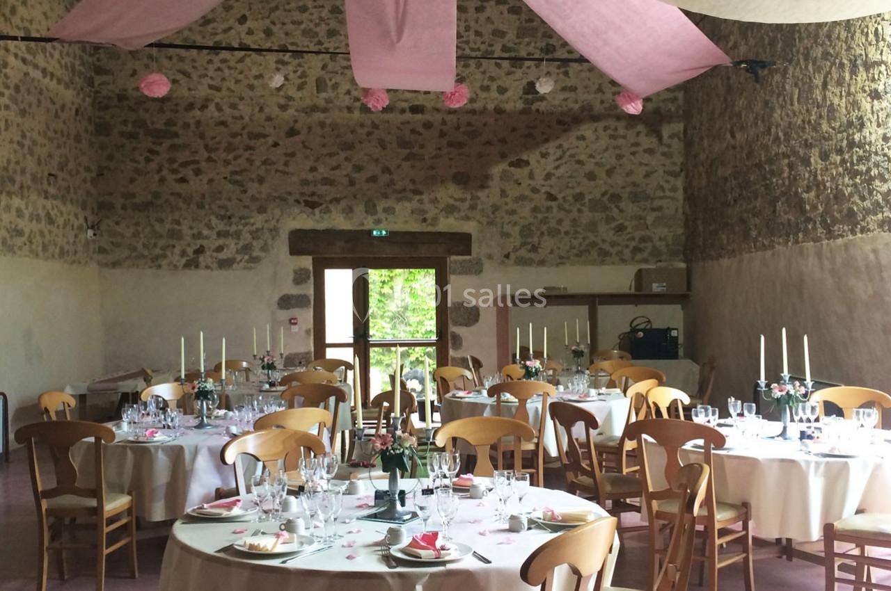 Salle de réception décorée pour un événement, avec tables rondes dressées, nappes blanches et guirlandes roses suspendues.