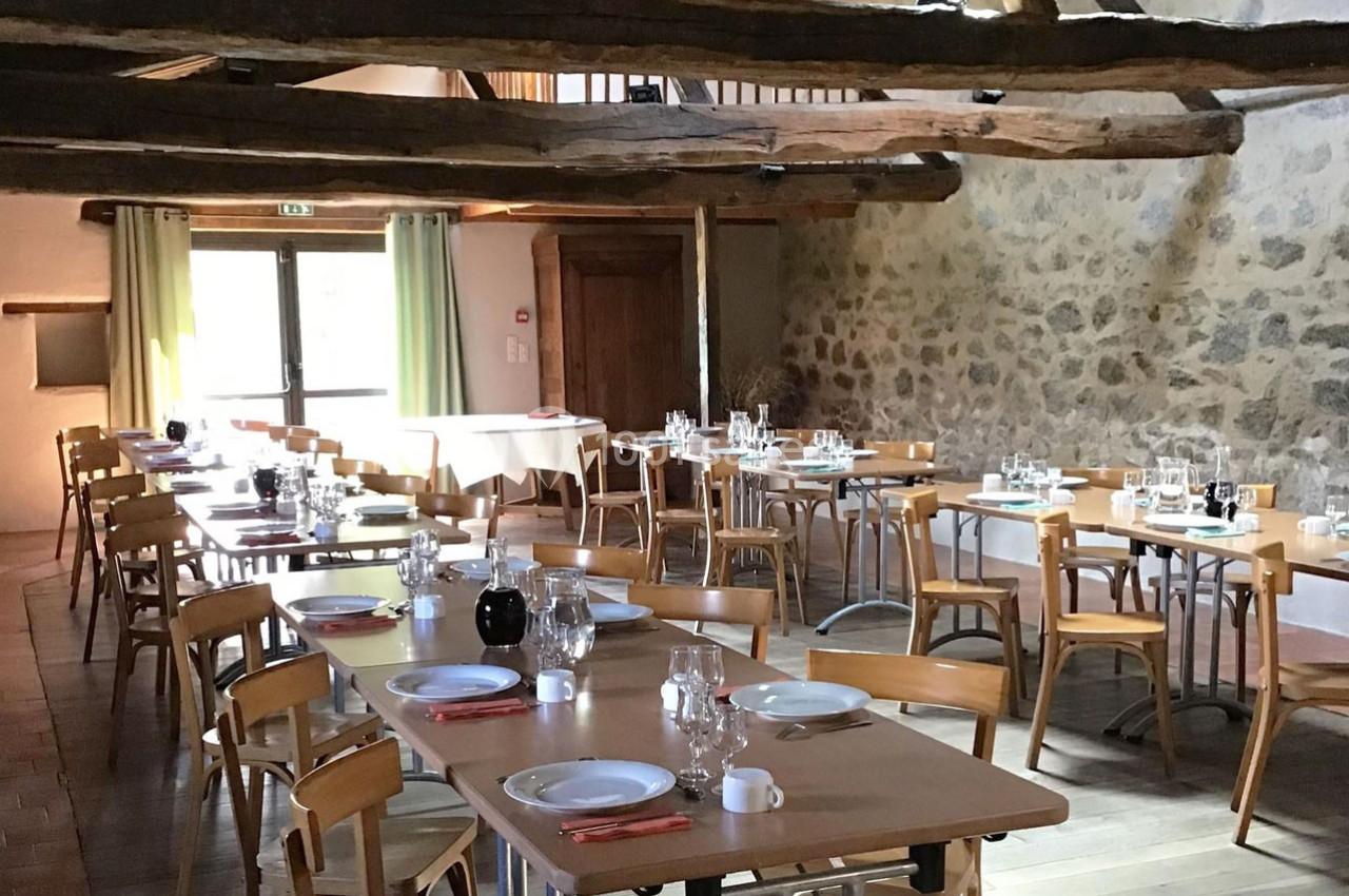 Salle rustique avec tables en bois dressées, vaisselle et poutres apparentes, éclairée par une lumière naturelle.